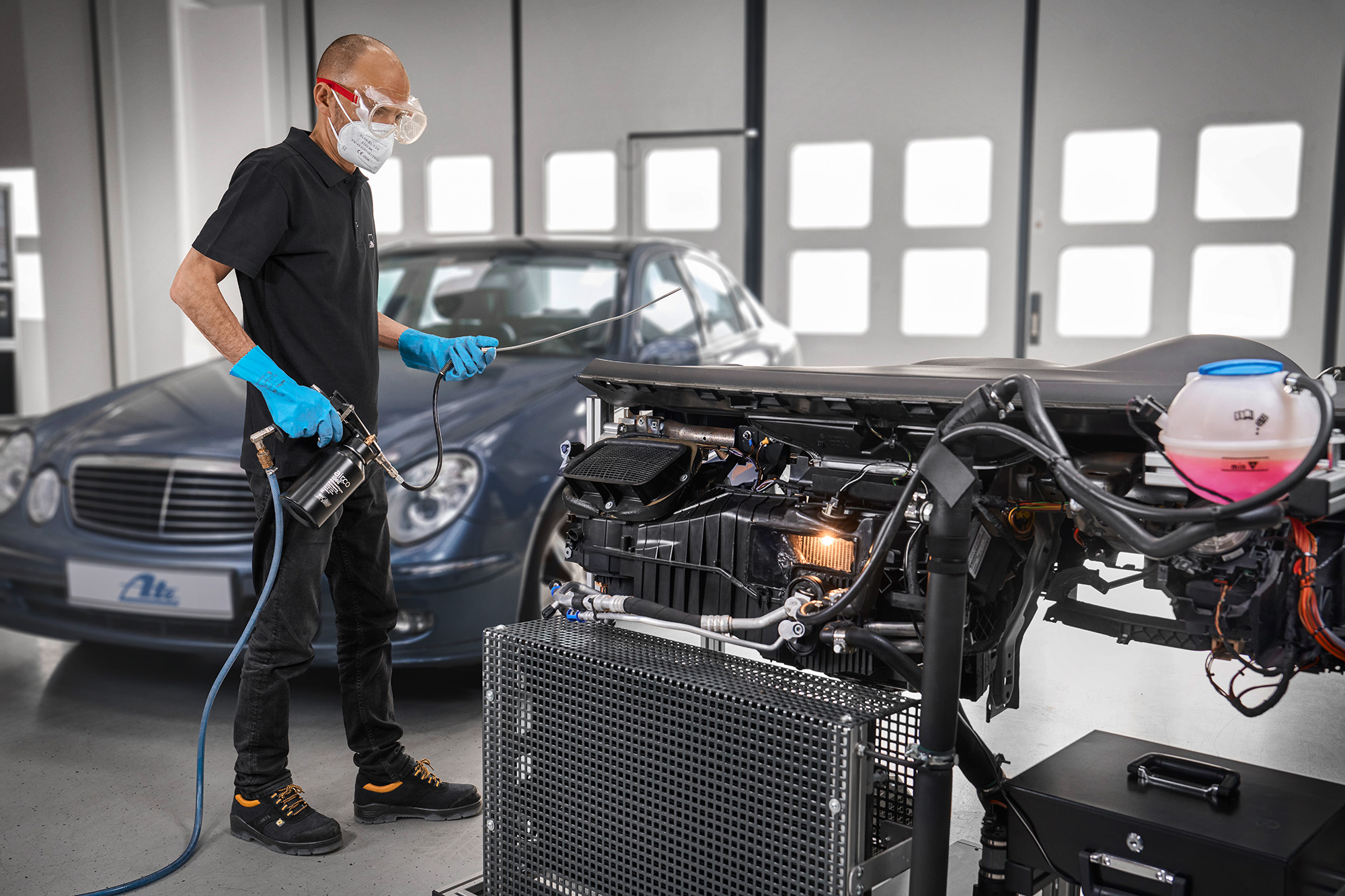 Bild in der Werkstatt, Person mit Maske, Schutzbrille, blauen Handschuhen bedient Gerätschaften, im Hintergrund steht ein Auto.